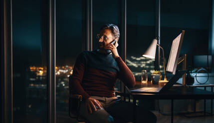 A dedicated businessman works late into the night at his desk, communicating with colleagues using his laptop and phone. The bustling office environment reflects a commitment to productivity and meeting deadlines. - JLPSF30789