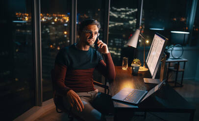 A dedicated business professional, working from home, multitasks during late night hours. Engrossed in a mobile phone call at his desk, he demonstrates commitment and focus. This image showcases remote work and work-life balance. - JLPSF30790