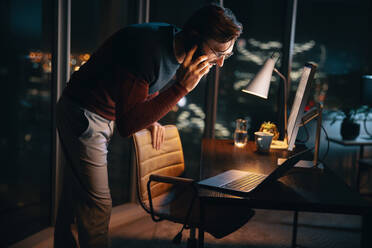 A business person working late at night in an office. He is discussing a project over a phone call while using a laptop and mobile phone. Dedicated and focused on meeting the deadline. - JLPSF30792