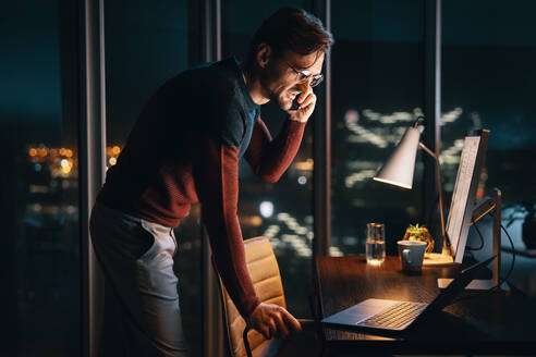 A dedicated business person, working late from a home office, discusses a project on a phone call. This caucasian male entrepreneur utilizes technology and remote work to ensure successful business operations. - JLPSF30793