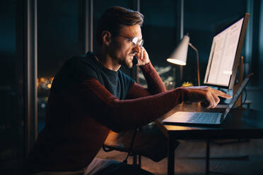A caucasian businessman works late at night in an office, discussing a project over the phone. He is focused and determined, balancing technology and communication for success. - JLPSF30794