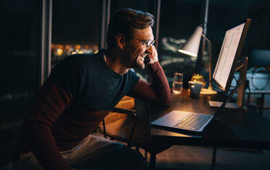 A happy business person works late at night in the office, discussing a project on a phone call. He sits at his desk, focused on his laptop, maintaining a professional work-life balance. - JLPSF30795