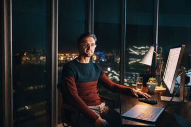 A caucasian male entrepreneur works late at his office desk, conducting market research with his laptop. He sits confidently, looking at the camera, showcasing dedication to his project. - JLPSF30796