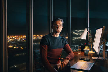 A focused Caucasian business man working late at night in an office. He sits at his desk, looking at the camera while diligently working on a laptop. This image captures dedication and commitment in the workplace. - JLPSF30797