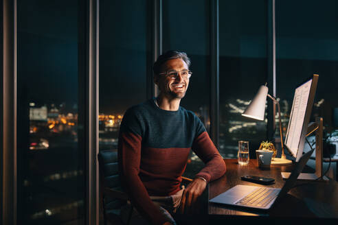 Happy caucasian business man sitting at his home office desk at night, working on a project with a smile on his face. Professional entrepreneur using technology and embracing remote work. - JLPSF30798