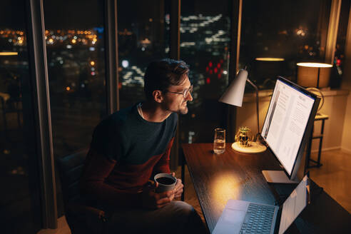 Ein Geschäftsmann arbeitet bis spät in die Nacht in seinem Büro zu Hause, um ein Projekt zu planen und zu leiten. Ein professioneller Mann sitzt konzentriert und engagiert an seinem Schreibtisch, schaut auf seinen Computerbildschirm und hält eine Tasse Kaffee in der Hand. - JLPSF30799