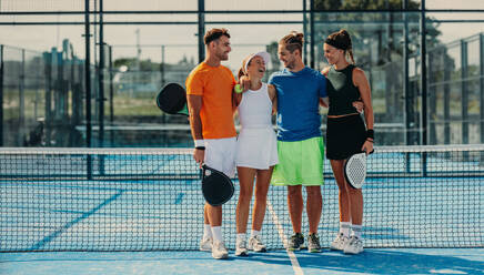 Ein Team von Männern und Frauen steht zusammen auf dem Padelplatz und lächelt glücklich, nachdem sie ein Wettbewerbsspiel für ihren Verein gewonnen haben. Eine Gruppe von Padelsportlern feiert den Sieg. Bei diesem Foto wurde absichtlich 35-mm-Filmkorn verwendet. - JLPPF02357