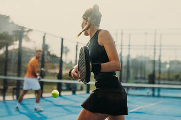 A female athlete stands on a padel court, receiving a shot with her padel racket. She wears sports clothing and competes in a tournament, displaying her skill in the game. This photo has intentional use of 35mm film grain. - JLPPF02358