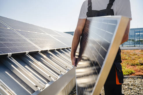 Handwerker, der ein Solarmodul auf dem Dach eines Firmengebäudes hält, aus einer Nahaufnahme betrachtet - DIGF20254