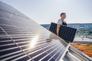 Ein Handwerker transportiert vorsichtig ein Solarmodul zur Installation auf das Dach eines Geschäftsgebäudes - DIGF20255