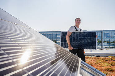 Ein Handwerker transportiert vorsichtig ein Solarmodul zur Installation auf das Dach eines Geschäftsgebäudes - DIGF20256