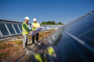 Zwei Techniker analysieren Pläne auf dem Dach eines mit Sonnenkollektoren ausgestatteten Firmengebäudes - DIGF20266