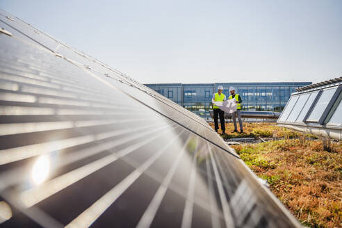 Two technicians analyzing blueprints on the rooftop of a corporate building equipped with solar panels - DIGF20269