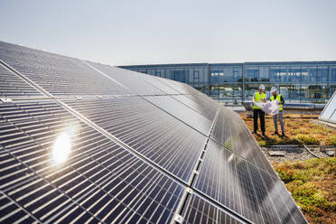 Zwei Techniker analysieren Pläne auf dem Dach eines mit Sonnenkollektoren ausgestatteten Firmengebäudes - DIGF20270