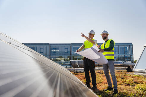 Two technicians inspecting solar panels on the roof of a company building, one pointing to a specific area - DIGF20271
