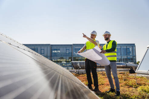 Zwei Techniker inspizieren Solarmodule auf dem Dach eines Firmengebäudes, einer zeigt auf einen bestimmten Bereich - DIGF20271