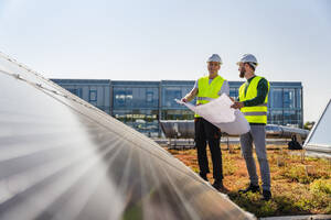 Two technicians analyzing blueprints on the rooftop of a corporate building equipped with solar panels - DIGF20272