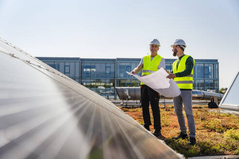 Two technicians analyzing blueprints on the rooftop of a corporate building equipped with solar panels - DIGF20272