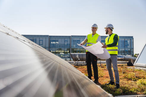 Zwei Techniker analysieren Pläne auf dem Dach eines mit Sonnenkollektoren ausgestatteten Firmengebäudes - DIGF20272