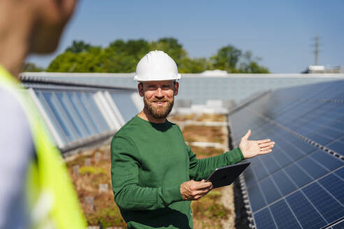 A man discusses business on a rooftop adorned with solar panels, using a tablet PC to communicate with a colleague - DIGF20280