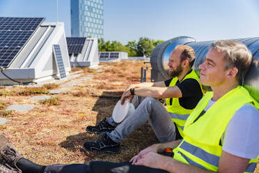 Zwei Techniker genießen eine Pause auf dem Dach eines Firmengebäudes, umgeben von Sonnenkollektoren - DIGF20290
