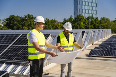 Zwei Techniker, die auf dem Dach eines mit Solarzellen bestückten Firmengebäudes Strategien entwickeln - DIGF20293