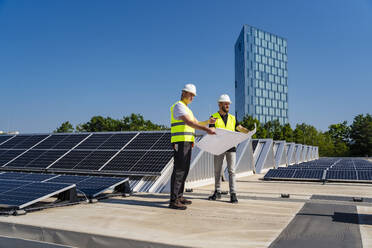 Zwei Techniker, die auf dem Dach eines mit Solarzellen bestückten Firmengebäudes Strategien entwickeln - DIGF20298