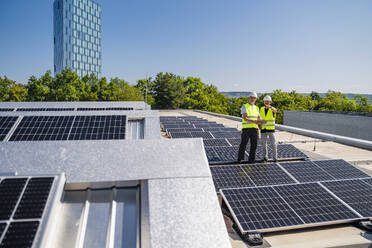 Zwei Techniker arbeiten mit einem Tablet-PC auf dem Dach eines Firmengebäudes, das mit Solarzellen ausgestattet ist - DIGF20323