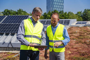 Two workers in safety vests discuss on the rooftop of a solar-powered company building, using a tablet computer - DIGF20332