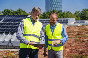 Zwei Arbeiter in Sicherheitswesten diskutieren auf dem Dach eines solarbetriebenen Firmengebäudes mit einem Tablet-Computer - DIGF20332