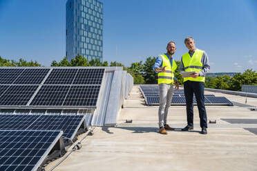 Zwei Arbeiter in Sicherheitswesten diskutieren auf dem Dach eines solarbetriebenen Firmengebäudes, während sie ein Tablet halten - DIGF20334