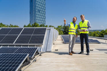 Zwei Arbeiter in Sicherheitswesten diskutieren auf dem Dach eines solarbetriebenen Firmengebäudes, während sie ein Tablet halten - DIGF20335