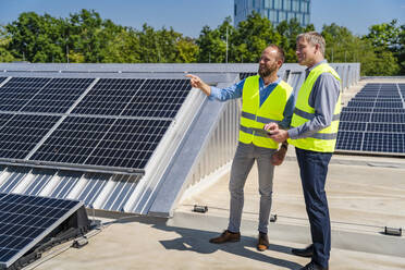 Zwei Arbeiter in Sicherheitswesten diskutieren auf einem Dach mit Solarzellen, während sie einen Tablet-Computer halten - DIGF20336