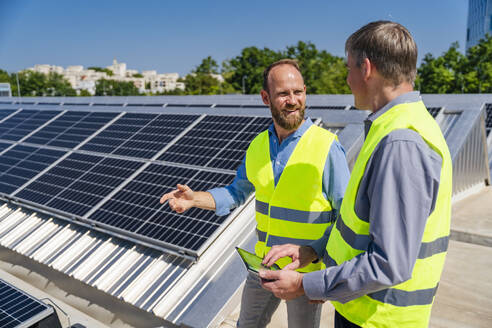 Zwei Arbeiter in Sicherheitswesten diskutieren auf dem Dach eines solarbetriebenen Firmengebäudes, während sie ein Tablet halten - DIGF20337