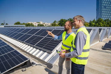 Zwei Arbeiter in Sicherheitswesten diskutieren auf einem Dach mit Solarzellen, während sie einen Tablet-Computer halten - DIGF20338