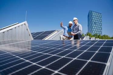 Zwei leitende Angestellte mit Schutzhelmen diskutieren auf dem Dach eines mit Solarzellen ausgestatteten Firmengebäudes - DIGF20341