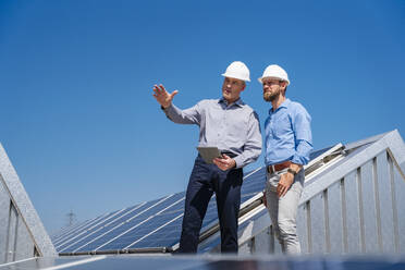 Zwei Geschäftsleute mit Schutzhelmen diskutieren über die Installation von Solarmodulen während eines Treffens auf dem Dach - DIGF20344