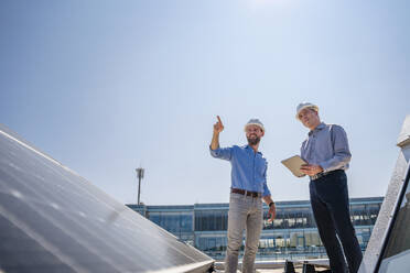 Zwei Führungskräfte in Schutzkleidung besprechen auf einem mit Sonnenkollektoren geschmückten Dach ihre Geschäfte - DIGF20353
