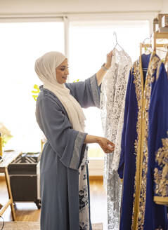 Lächelnde junge Modedesignerin bei der Auswahl eines Hochzeitskleides in einer Boutique - JCCMF10684