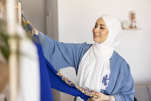 Glückliche junge Modedesignerin mit Hidschab, die ein blaues Hochzeitskleid betrachtet - JCCMF10688