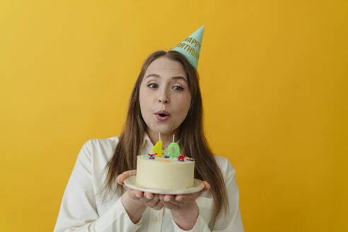 Woman wearing party hat holding birthday cake against yellow background - OSF02010