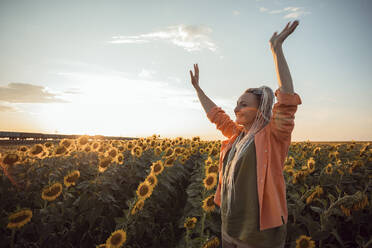 Lächelnde Frau mit erhobenen Armen in einem Sonnenblumenfeld stehend - ADF00145