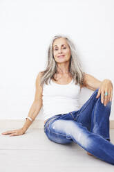Lächelnde Frau in Jeans, die zu Hause auf einem Hartholzboden sitzt - JBYF00246