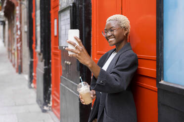 Smiling businesswoman taking selfie through smart phone in front of building - MMPF00823