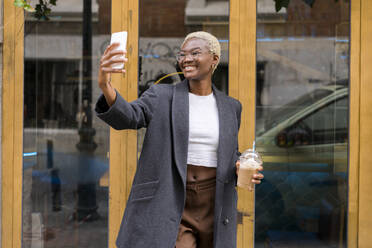 Happy businesswoman taking selfie through smart phone near glass door - MMPF00826