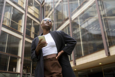 Contemplative businesswoman holding smart phone outside office building - MMPF00832