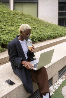 Junge Geschäftsfrau nippt an einem kalten Kaffee und benutzt einen Laptop im Büropark - MMPF00835