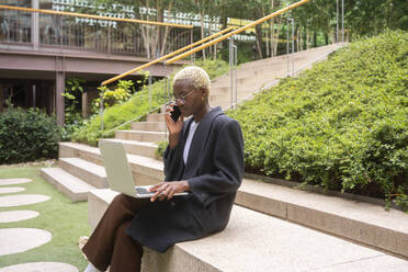 Geschäftsfrau, die einen Laptop hält und mit einem Smartphone vor einem Bürogebäude telefoniert - MMPF00836