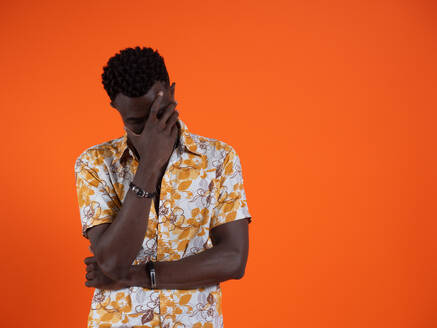 Depressed young African American male in orange printed short sleeves shirt and closed eyes while standing with crossed arms and hiding face in hand fingers against orange background - ADSF46636