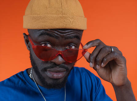 Stylish bearded African American male in hat adjusting red eyeglasses, blue shirt and neck chain looking at camera against orange background - ADSF46639
